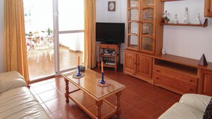 Een woonkamer met een salontafel met glazen blad, een televisie, houten kasten en een zichtbare eethoek door een glazen deur.