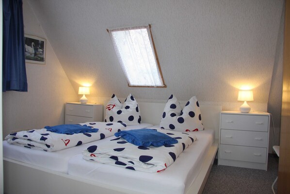 A twin bed with a white and blue polka dot bedspread, two bedside tables with lamps, a small window with lace curtains, and a wall-mounted picture.