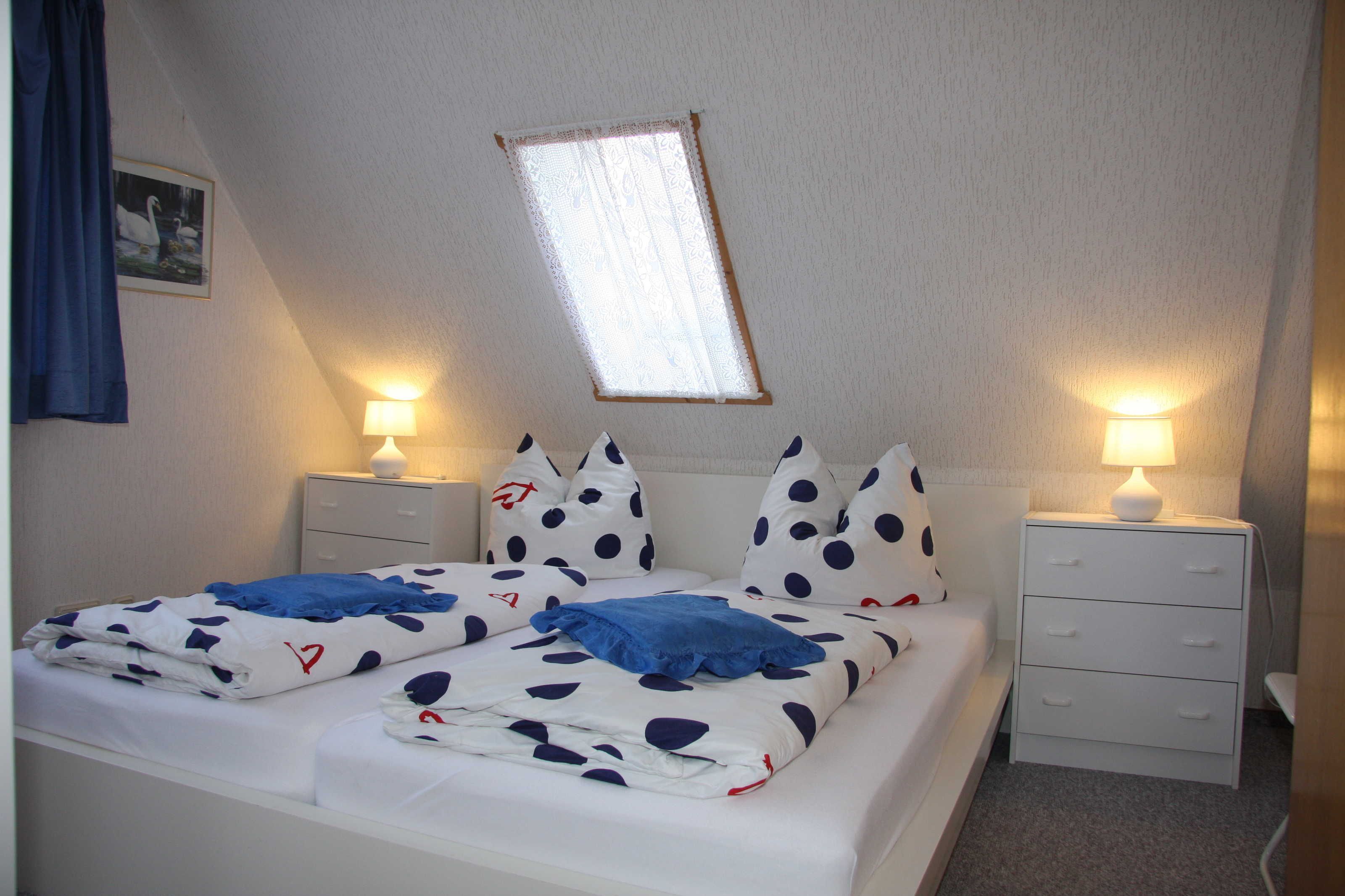 Ein Doppelbett mit weißer und blauer Punkteoptik, zwei Nachttische mit Lampen, ein kleines Fenster mit Spitzenvorhängen und ein an der Wand hängendes Bild.