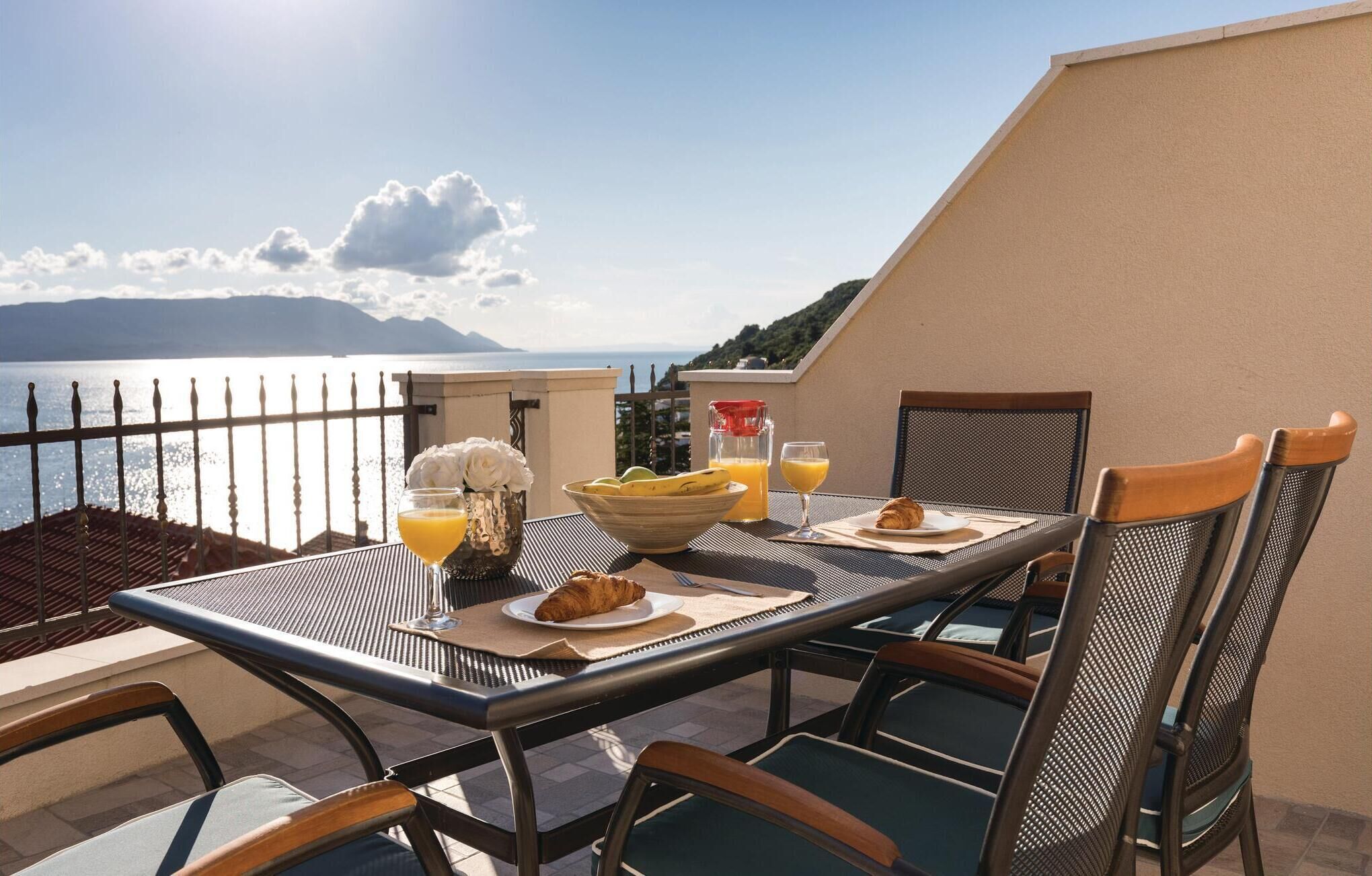 Ein Esstisch mit Stühlen, ein Croissant, eine Obstschale, ein Glas Orangensaft und eine Saftflasche auf einem Balkon mit Blick auf ein Gewässer und Berge.