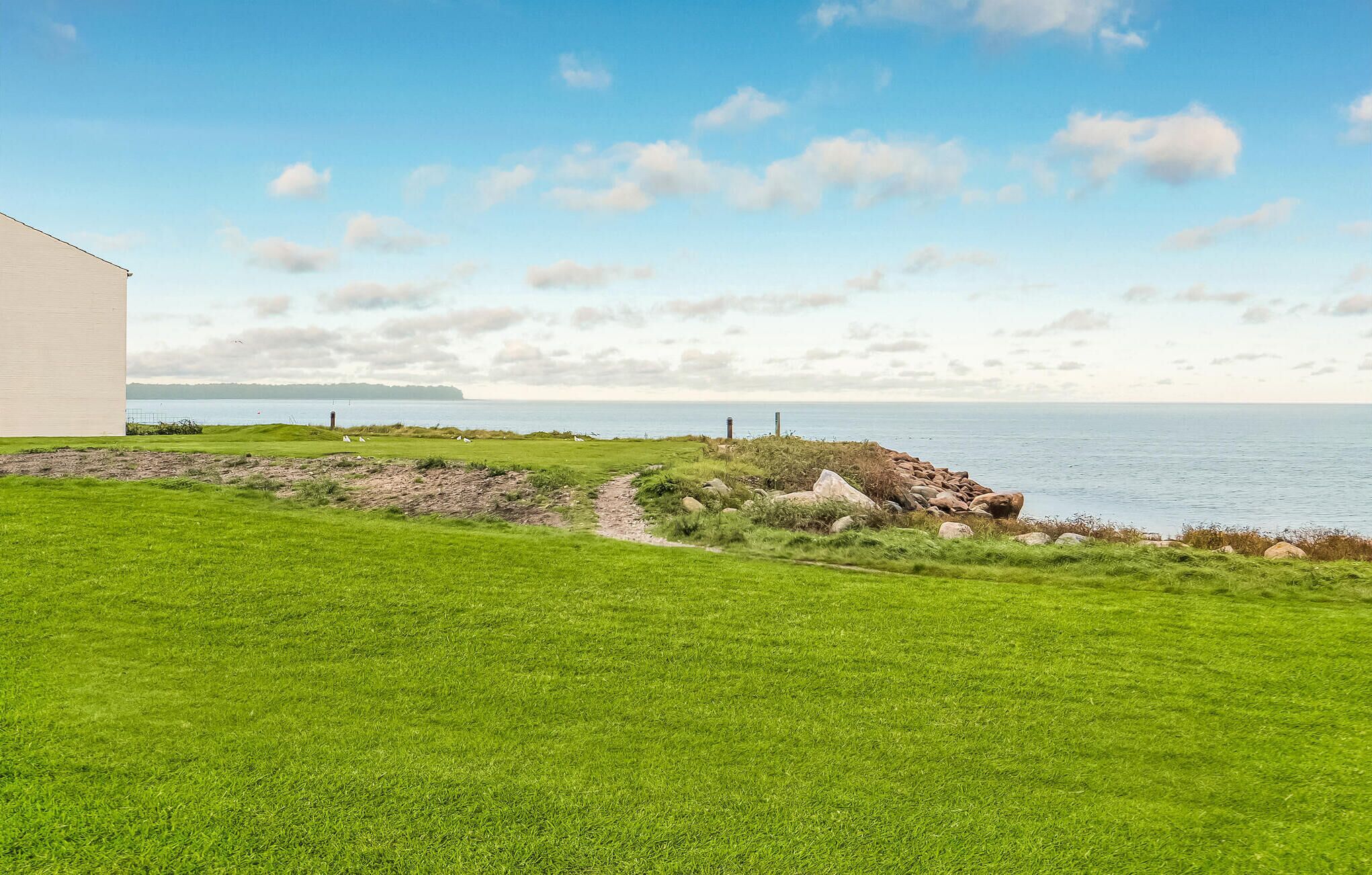 A grassy area with a path leading to a rocky shoreline by the sea, a white building, and a clear sky.