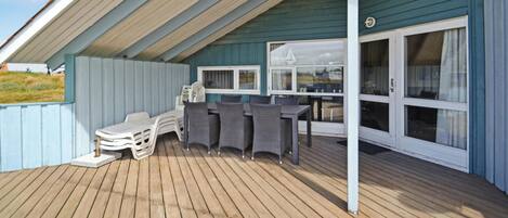 A covered outdoor area with a wooden deck, a table with chairs, and a window with curtains.