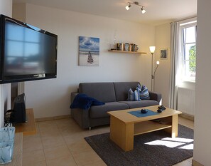 Una sala de estar con un televisor de pantalla plana, un sofá gris, una mesa de centro de madera y una estantería con libros.