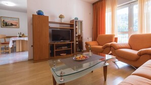 A living room with a glass coffee table, orange sofa, TV, wooden cabinet, and a dining area with a table set for two. - 3 bedroom gorgeous home in Duba (Duba)