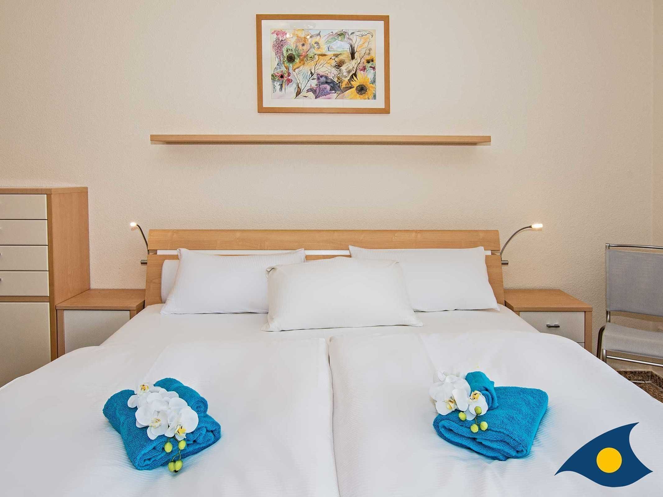 Un lit double bien fait, avec du linge de lit blanc, deux serviettes bleues ornées de motifs floraux, une tête de lit en bois et un tableau encadré au-dessus.