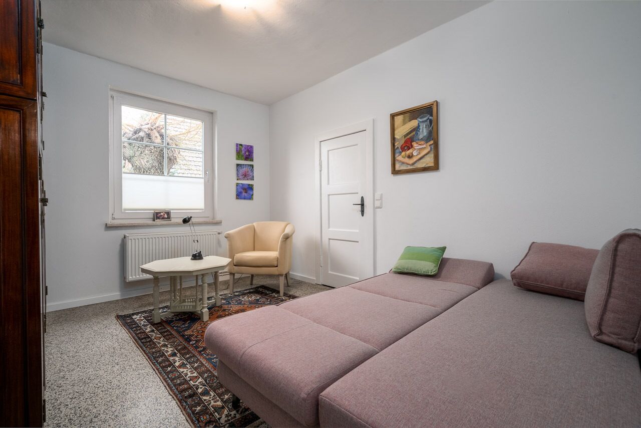 A bedroom with a bed, a chair, a table, a radiator, a window, and a door.
