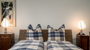 Uma cama de casal com colcha xadrez, duas almofadas combinando, cabeceira de vime e duas luminárias de cabeceira.