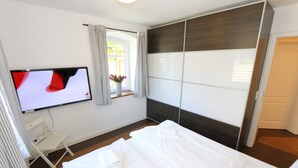A hotel room with a bed, a TV mounted on the wall, a window with curtains, and a bedside table with a vase of flowers.
