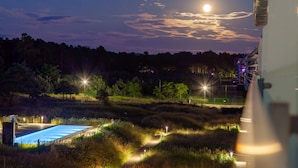 Vista nocturna de un área ajardinada con piscina, senderos y árboles iluminados.