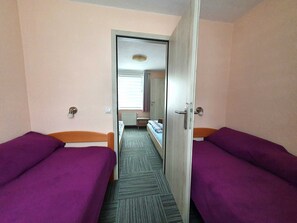 A narrow corridor leading to a room with two beds, each with a purple bedspread, a wooden headboard, and a small bedside table.