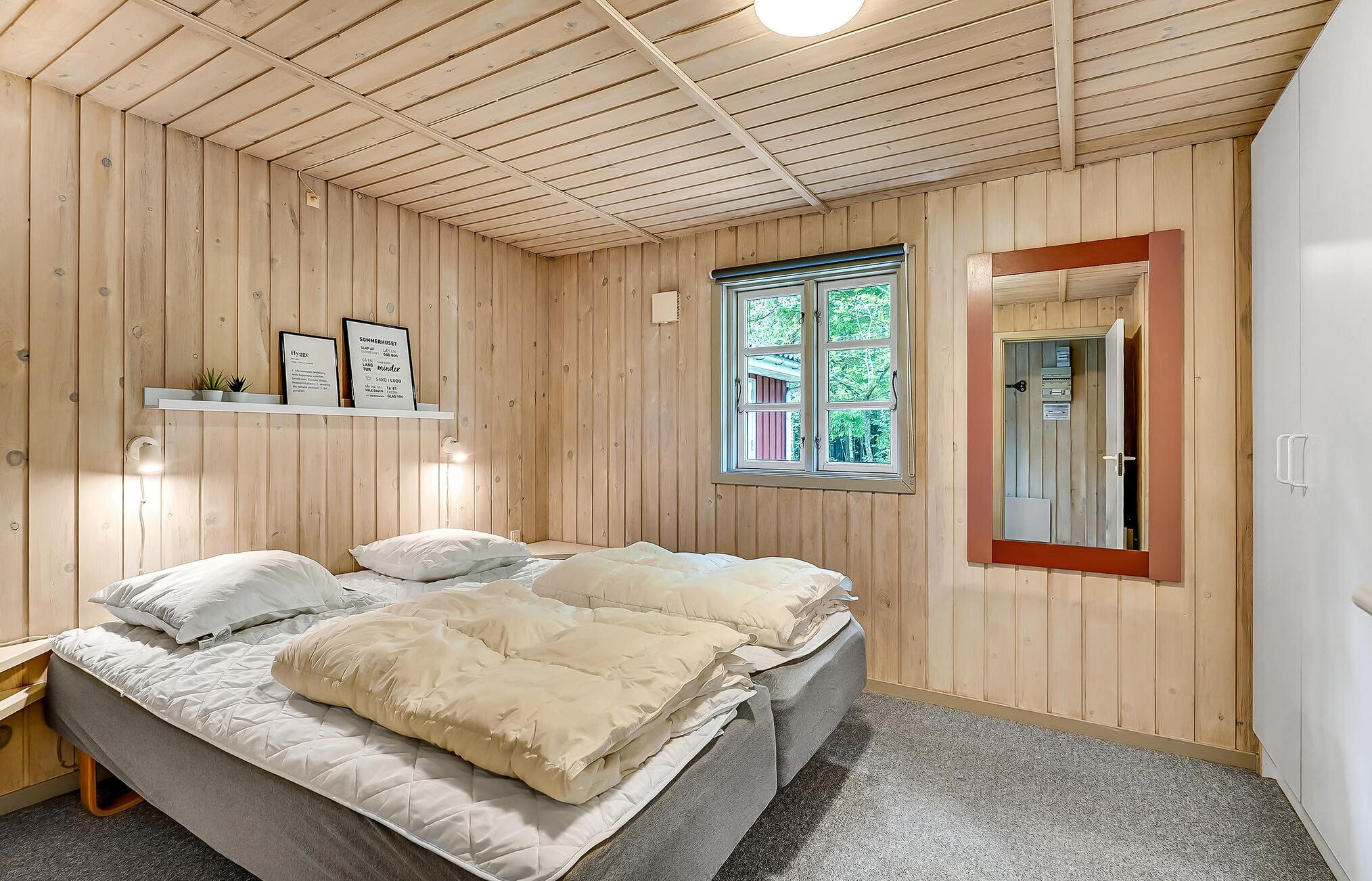 A bedroom with a wooden ceiling, a bed with white and beige bedding, a small shelf with framed pictures, a window with a view of greenery, and a door with a mirror.