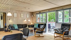 Una habitación acogedora con paredes y techo de madera, y ventanas amplias que ofrecen vistas a la vegetación exterior. Hay dos sofás, una mesa de comedor y dos sillones mecedores.