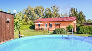 A swimming pool with a wooden deck, a small house with a red roof, and a wooden shed in a sunny outdoor setting.