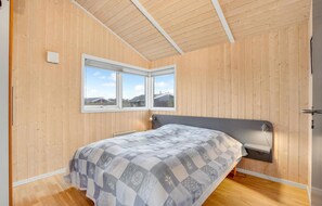 Een slaapkamer met houten wanden, een bed met een gedessineerd dekbedovertrek, een raam met uitzicht op huizen, en een plafond met houten balken.