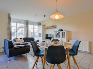 Ein modernes Wohnzimmer mit Essbereich, ausgestattet mit einer Sofagarnitur, einem Esstisch mit Stühlen, einem Fernseher und einer Schrankwand mit Regalen.