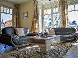 A modern living room with two black leather sofas, a wooden coffee table, and a patterned rug. Large windows with curtains allow natural light and views of buildings outside.