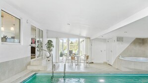A swimming pool with a clear blue water, surrounded by white walls and a tiled floor. There are chairs and a table in the background, and a window on the left side. - Awesome home in Væggerløse with sauna (Væggerløse)