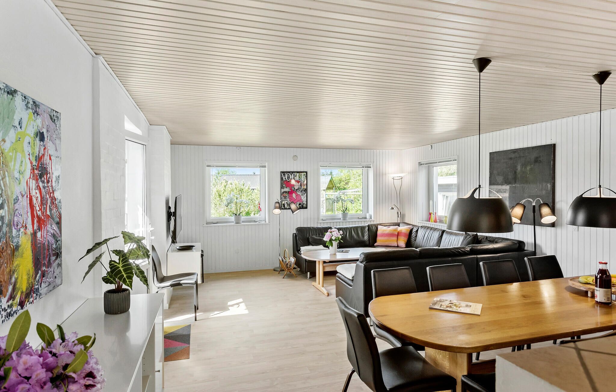 A modern interior with a wooden dining table, black leather chairs, a black leather sofa, and a white wall with a large abstract painting.