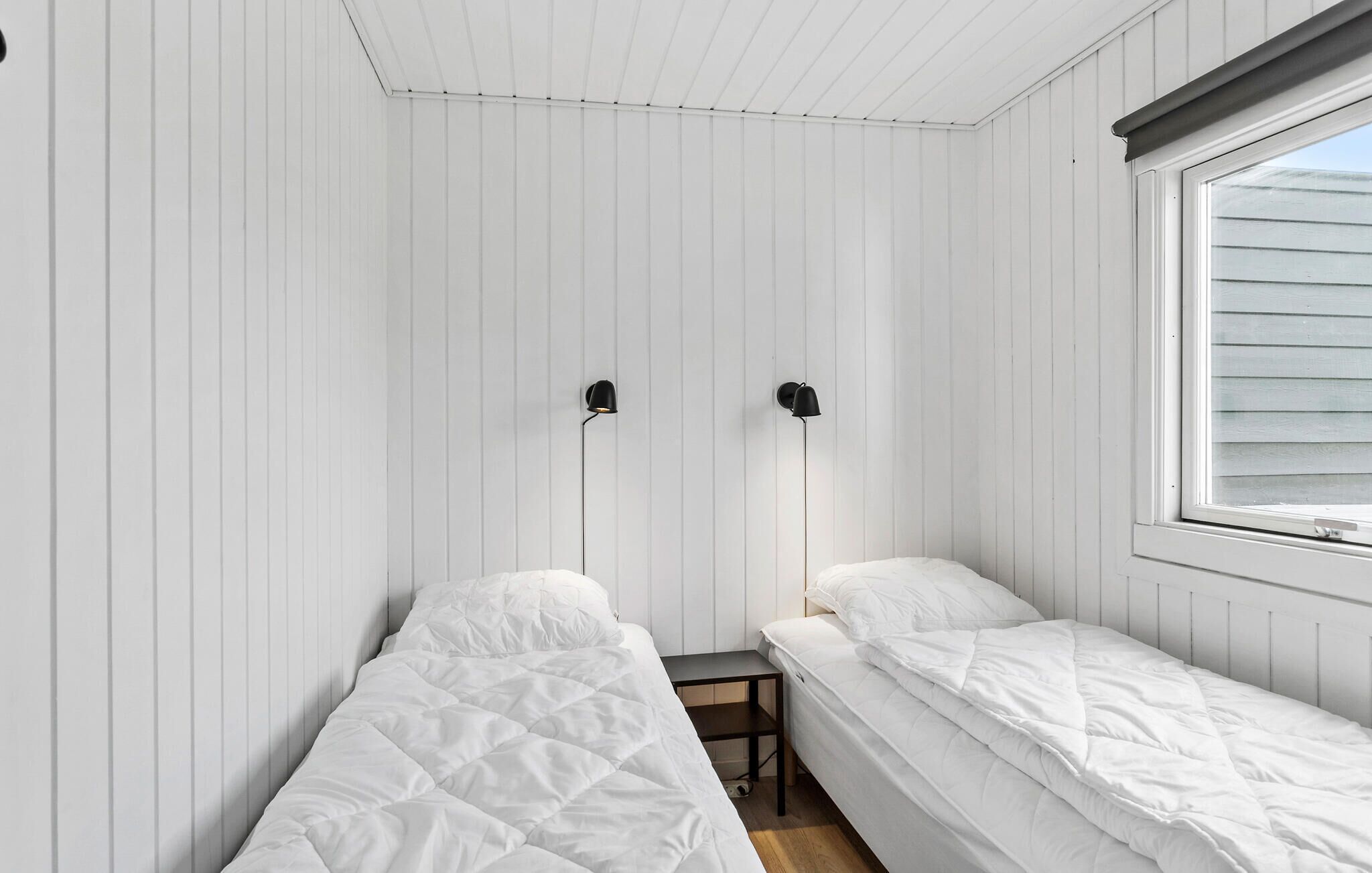 A small, single-room bedroom with two beds, a window, and two wall-mounted lamps.