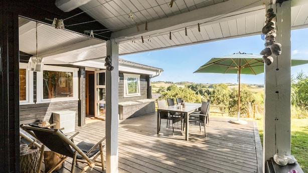 Une terrasse extérieure couverte, avec une table et des chaises, une maison en bois en arrière-plan et une vue sur la campagne.