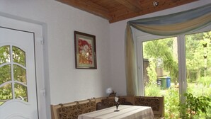 A room with a wooden bench, a table with a tablecloth, a framed picture on the wall, and a window with a view of greenery. - Flower House - Flower House * (Rankwitz)