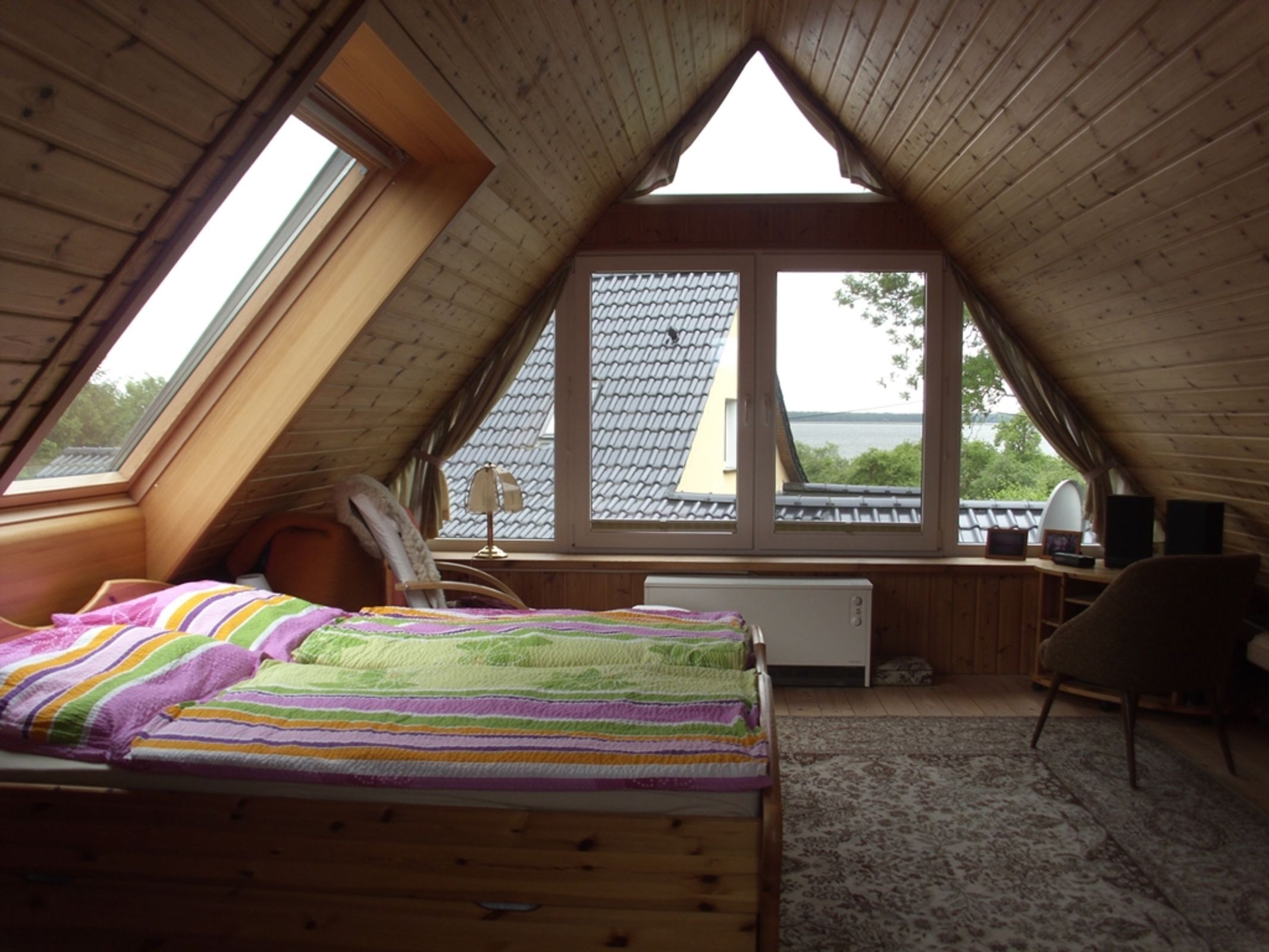 Une chambre avec un plafond en pente, un lit recouvert d’une couverture à rayures colorées, un bureau avec une lampe et une vue sur l’extérieur.