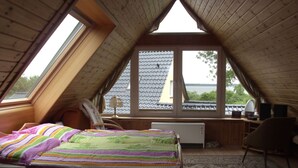 A bedroom with a sloped ceiling, a bed with a colorful striped blanket, a desk with a lamp, and a view of the outside. - Flower House - Flower House * (Rankwitz)