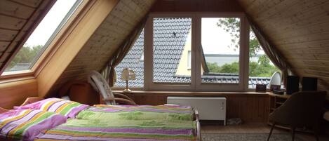 Une chambre avec un plafond en pente, un lit recouvert d’une couverture à rayures colorées, un bureau avec une lampe et une vue sur l’extérieur.