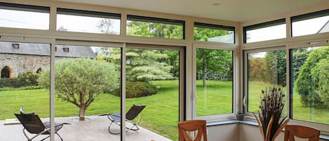 A dining area with a table and chairs, large windows, and a view of a garden and stone building.