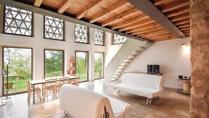 A spacious room with a high ceiling, large windows, a wooden staircase, a dining area, and two beds covered with white sheets.