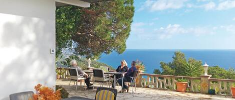 An outdoor seating area with stone flooring, tables, and chairs, overlooking a scenic view of the sea.
