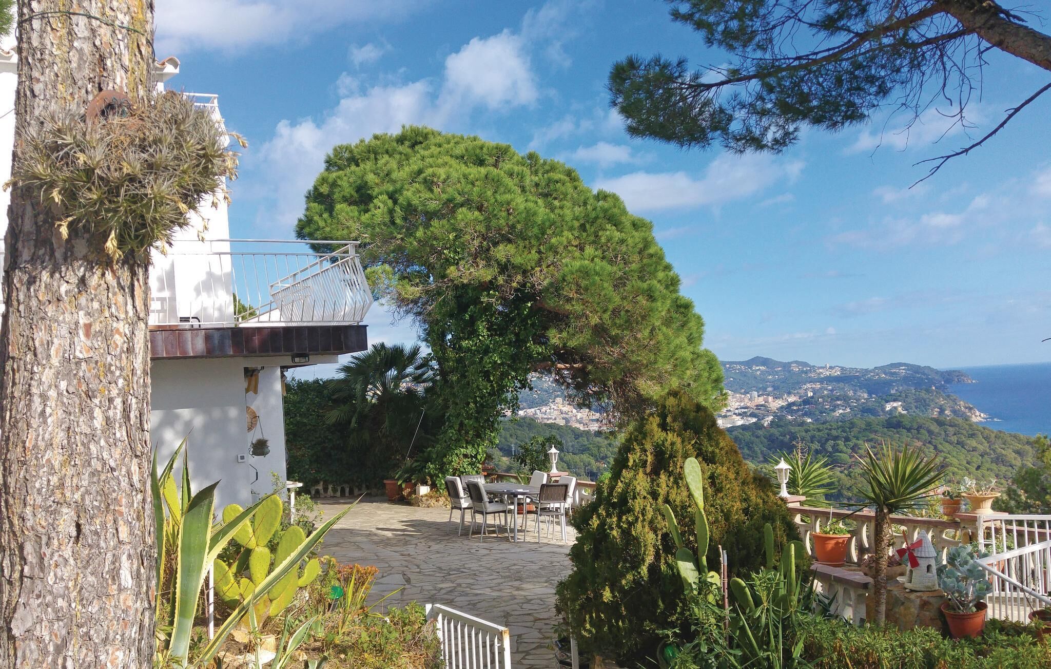 Un sendero de piedra que lleva a un edificio blanco con balcón, rodeado de abundante vegetación y vistas a un pueblo costero.