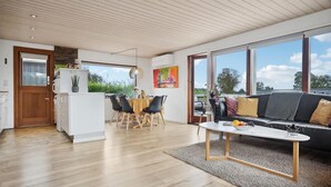 Amplia sala de estar con sofá, mesa de comedor y sillas. Zona de cocina con refrigerador y una mesa de comedor. Ventanas amplias ofrecen vistas al exterior.