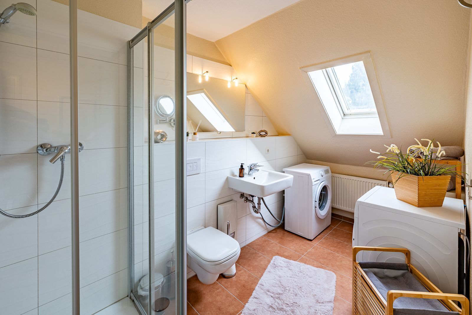 A compact, modern bathroom with a shower, toilet, sink, and washing machine. It features a skylight and a small window, providing natural light.