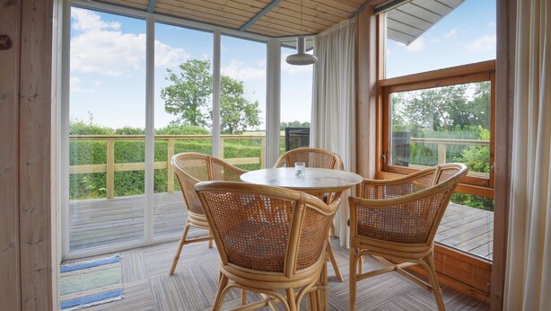A covered patio with a round table and four wicker chairs, large windows offering a view of greenery, and a wooden ceiling. - Nice home in Hejls with WiFi (Hejls)