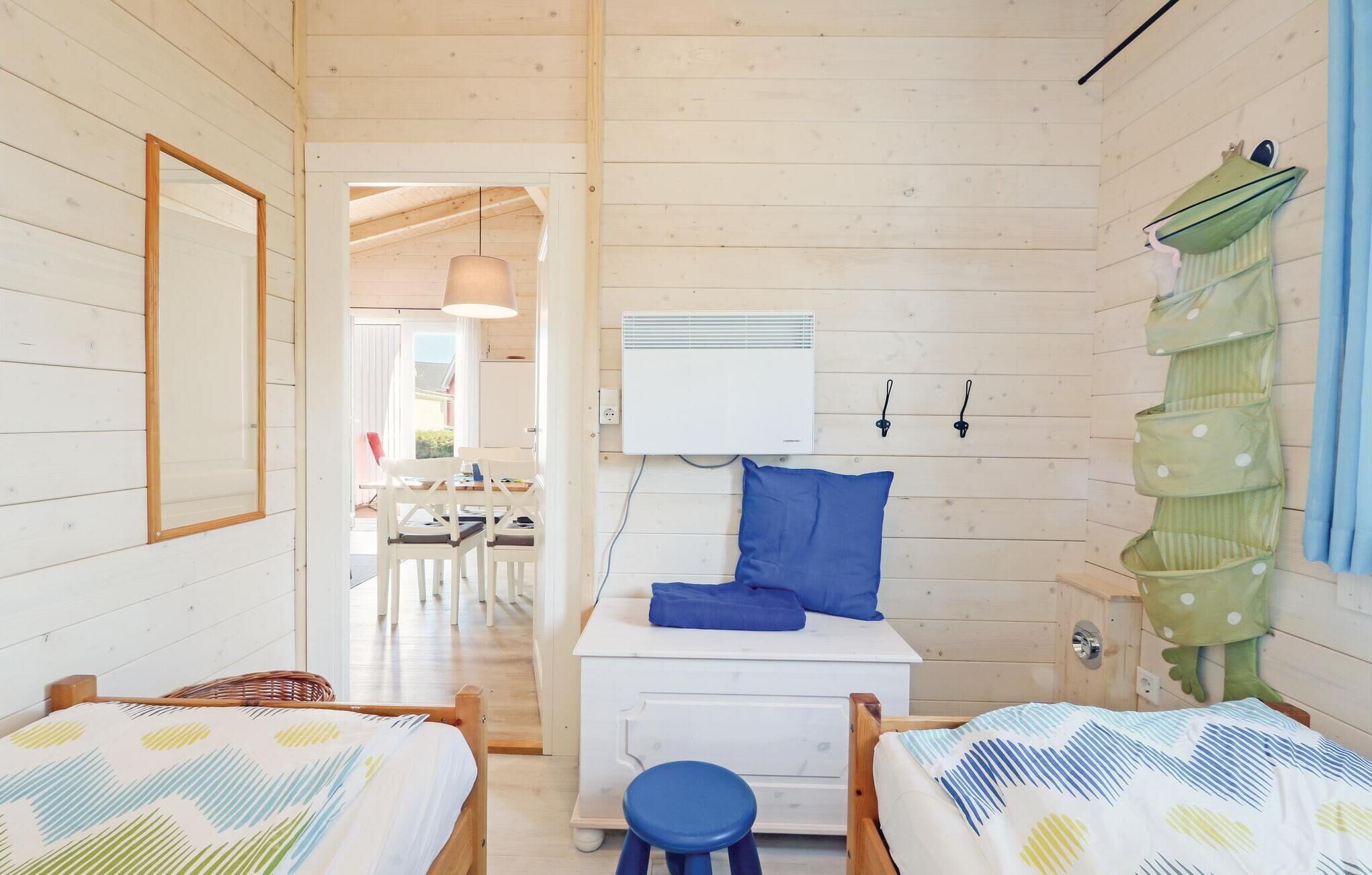 Quarto aconchegante com duas camas, uma mesinha e uma cadeira. Possui uma janela com cortinas, um aquecedor fixado na parede e um móvel de armazenamento verde.