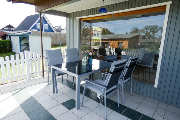 Een overdekt terras met een glazen tafel en stoelen, een wit hek en uitzicht op een huis en tuin.