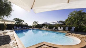 Ein Swimmingpool mit Liegestühlen und Sonnenschirmen, umgeben von Steinplatten und Grünpflanzen.