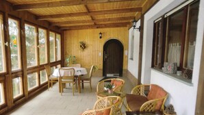Ein Zimmer mit Holzwänden, das eine Essecke mit Korbstühlen und einem Tisch, große Fenster und eine Tür mit einem Kranz bietet.
