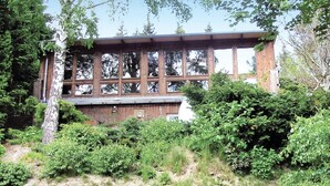 Eine Holzhütte mit großen Fenstern, umgeben von dichtem Grün und Bäumen.