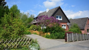 Een tweelaags huis met een schuin dak, omringd door een houten hek, en een geplaveid opritje dat naar de voordeur leidt.