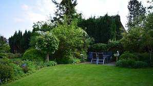 Een verzorgde tuin met gazon, diverse struiken en bomen. Er is een zitgedeelte met stoelen en een tafel.