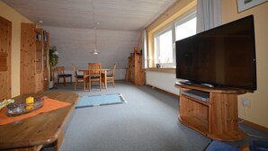 Een woonkamer met een houten tv-meubel, een eettafel met stoelen, een groot raam en een plafondventilator.