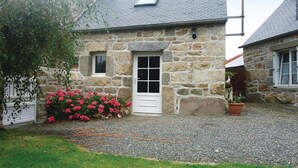 Een stenen huis met een witte deur, een klein raam en een tuin met roze bloemen.