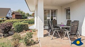 A covered patio with a table and chairs, a white brick wall, and a garden with bushes and a small tree. - Apartment (ap.) Whg. 01 Gartenstraße - Garden 01 (Heringsdorf)