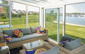 A sunlit room with a large glass door leading to a grassy area and a lake. Inside, there is a wicker sofa with colorful cushions and a glass coffee table. - Porta Isola - Villa Water 105 (Stevensweert)