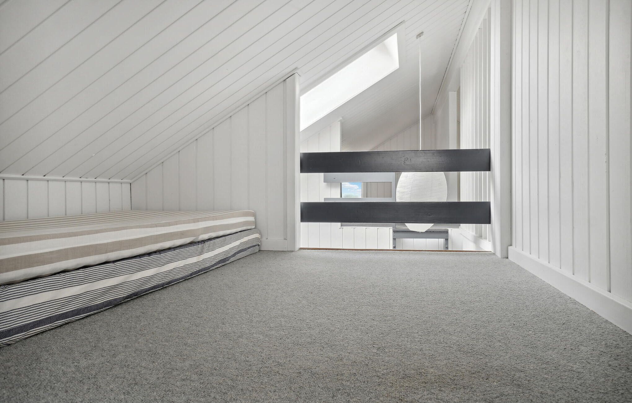 A high-ceilinged, white-walled room with a sloped ceiling, a bed with a striped mattress, and a bench.