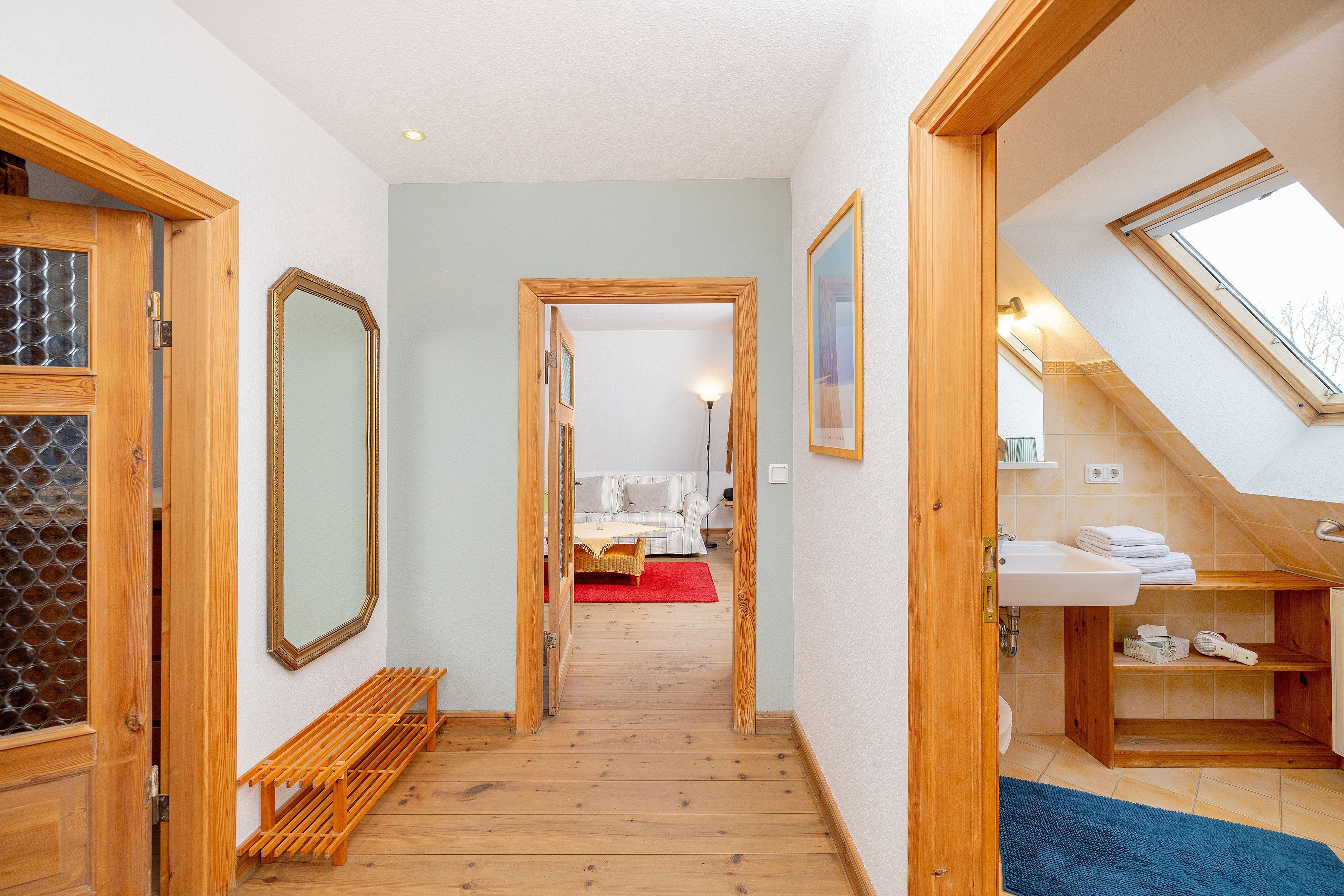 Ein Flur, der zu einem Schlafzimmer mit Dachfenster, einer Holzbank und einem Badezimmer mit Waschbecken und Handtuchhalter führt.