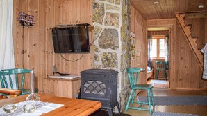Interior acogedor de una cabaña con pared de piedra, paredes de madera, televisor montado en la pared, mesa de madera con sillas y chimenea.