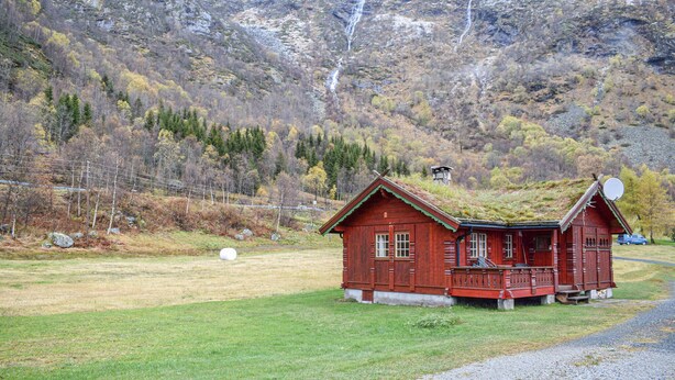 Una cabaña de madera roja con techo verde, rodeada por un campo cubierto de pasto y montañas al fondo.
